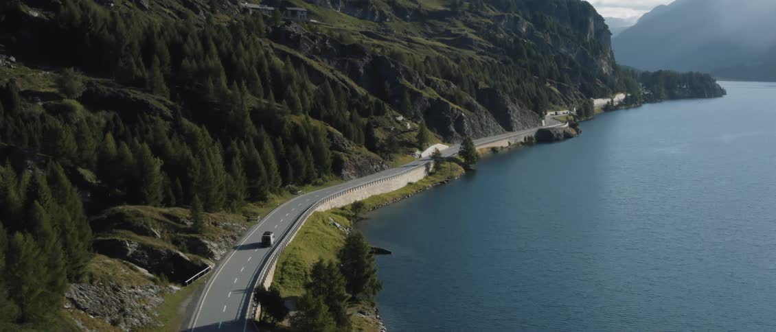 A car travels along a winding mountain road situated between a steep cliffside and a large blue lake