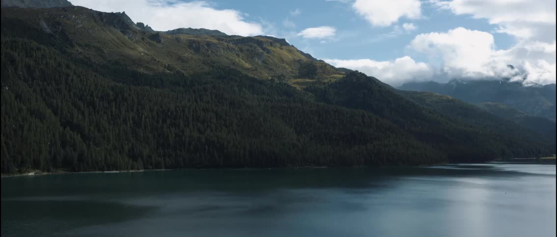 A vast calm body of water fills the foreground and reflects the soft light of a cloudy skysteep mountain slopes covered in dense evergreen forests rise directly from the shoreline toward rocky sundrenched peaks