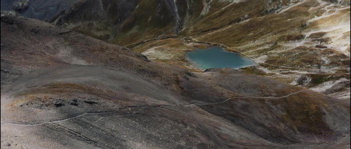 A mountainbiker rides a trail, in the background rests  a pond beneath a mountain ridge in the wilderness