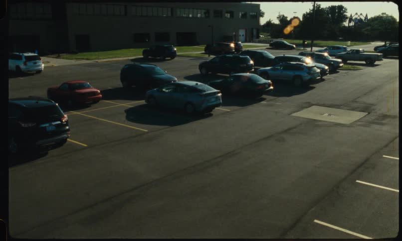 A highangle view of several vehicles parked in a sunlit asphalt parking lot during the day the scene captures a dark car pulling into an empty parking space while the sun creates a bright lens flare in the upper right corner