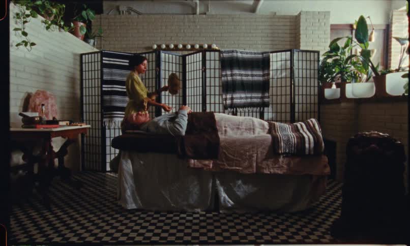 A robed practitioner moves around a person resting on a covered table gently brushing and hovering their hands over the client before stepping to a side table with small items in a calm indoor spalike room
