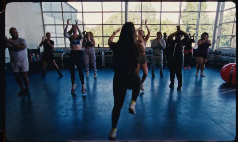 A lively group follows an energetic instructor in a dance fitness class inside a bright gym with large windows