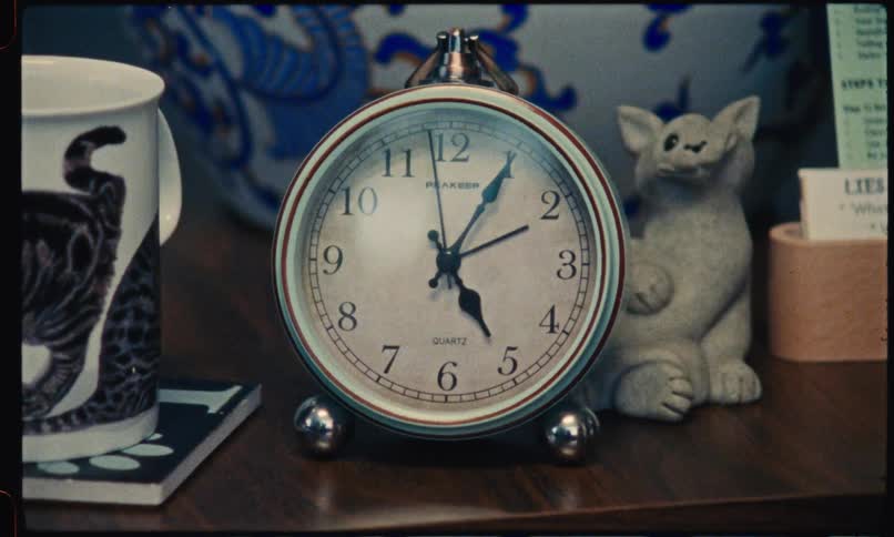 An analog alarm clock sits on a wooden shelf next to a mug and figurines as the camera zooms in