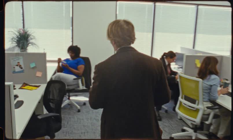 A man walks into an office workspace and sits down at his cubicle