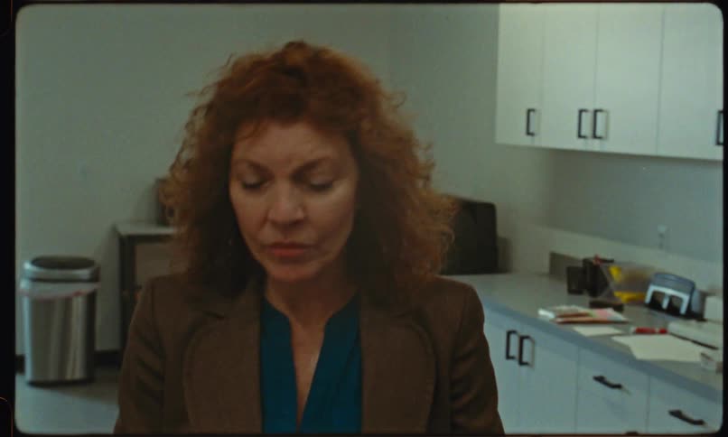 A woman wearing a green blazer with a blue top retrieves several documents from the printer in the office and walks out becoming shocked as she heads away