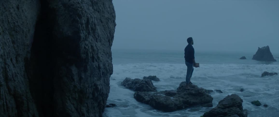 A man stands near a large rock formation while looking out at the ocean