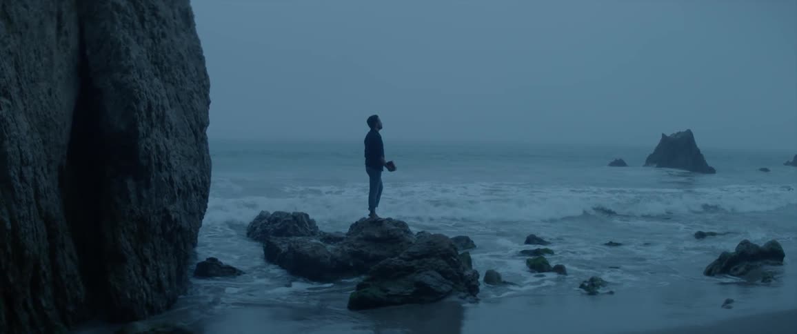 Man standing on rocks on beach as ocean waves crash in on foggy evening