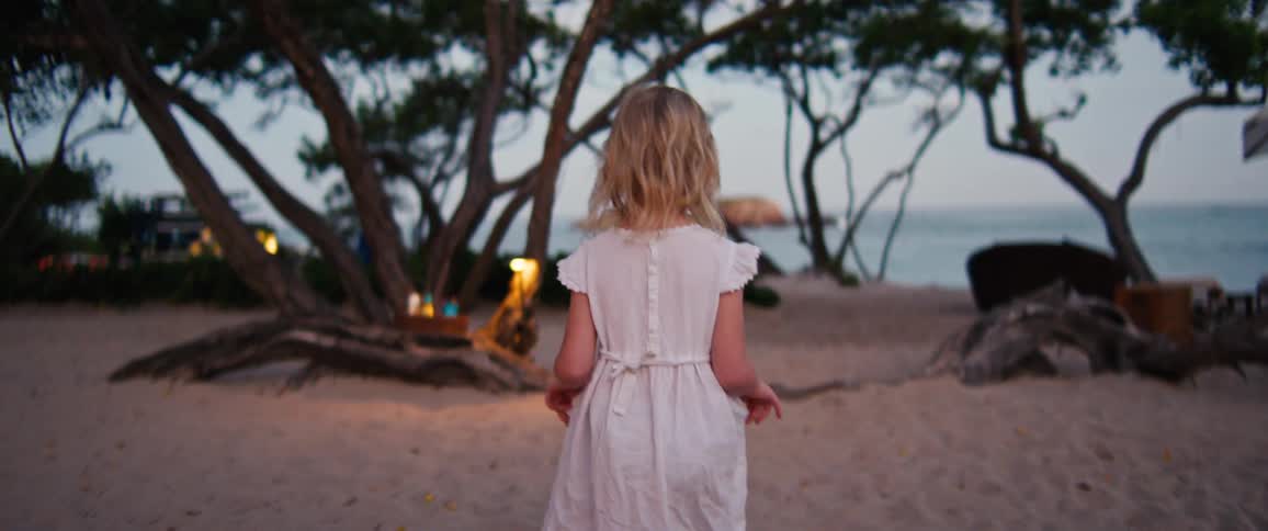 Young blonde girl wearing a white dress walking on a camping site next to the ocean outdoors during sunset time