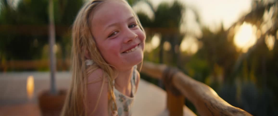 A young blonde girl smiles and laughs while leaning against a wooden railing on a balcony during sunset with tropical trees visible in the soft background