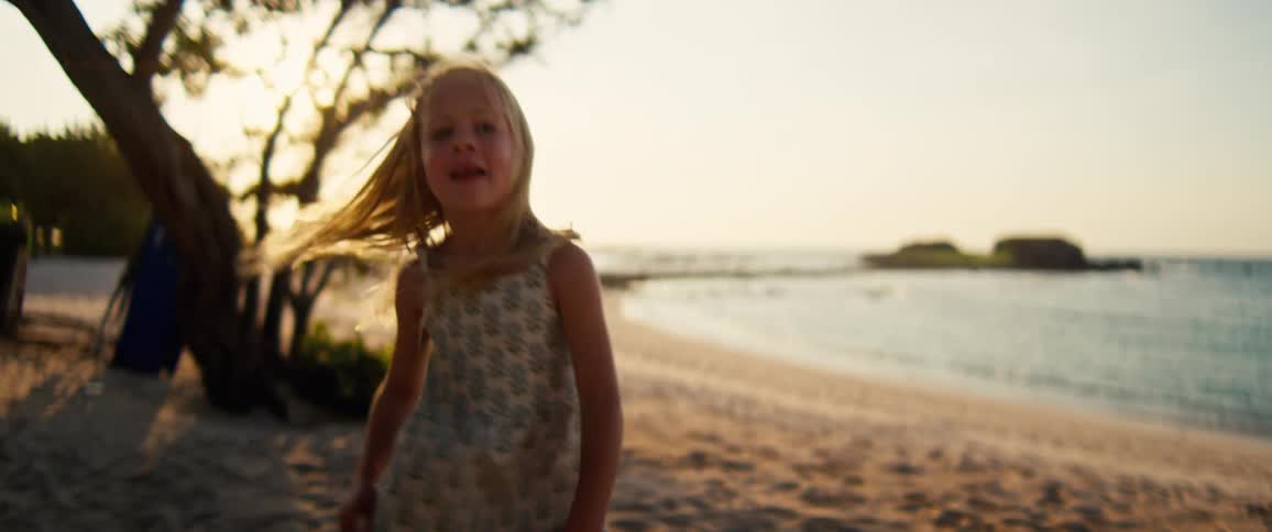 A young girl wearing a floral dress spins and dances on a sandy beach during sunset with the ocean and small islands in the background