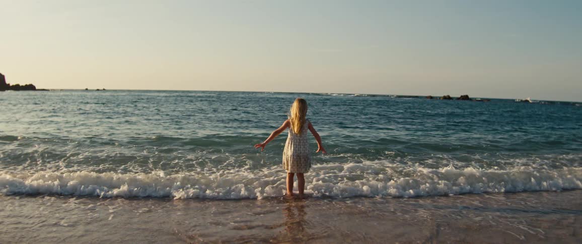A young girl stands and walks at the edge of the ocean with her arms outstretched as gentle waves roll in around her feet under a clear sky