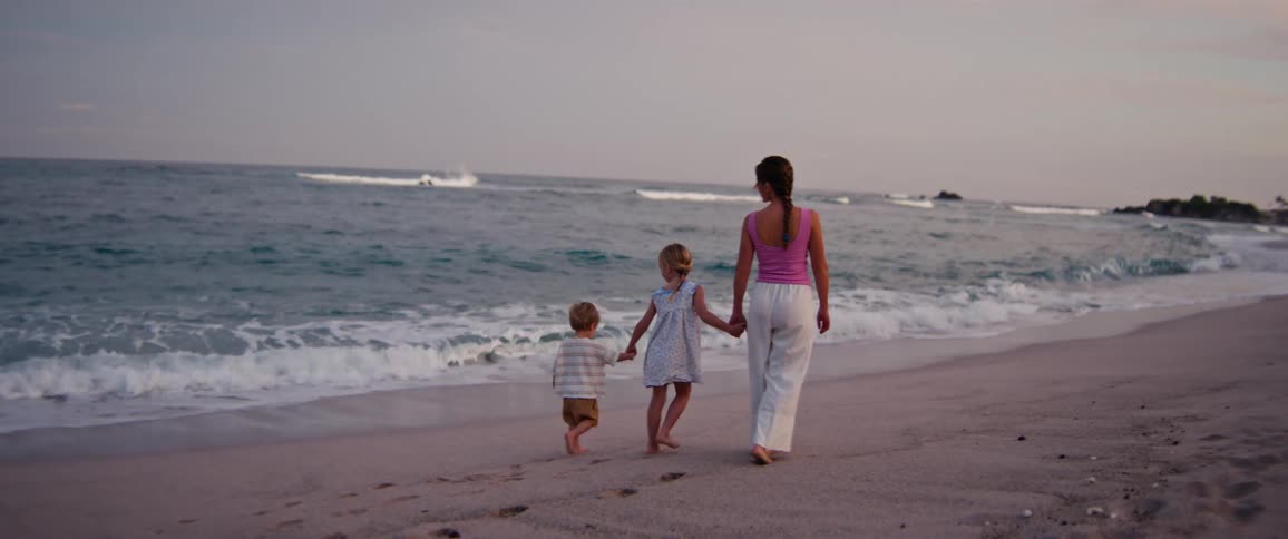 A woman walks hand in hand with two young children along the shoreline as waves gently roll onto the beach at sunset