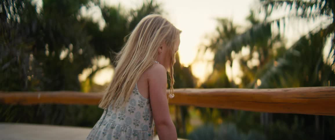 Blonde girl leaning on a wooden railing outdoors with palm trees in the background