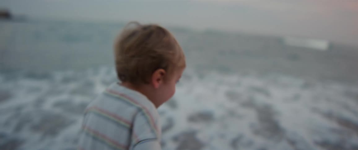 Little boy running barefoot on the beach as waves roll in to shore and over the sand as his mother picks him up and carries him away