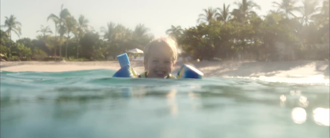 A young boy wearing blue floaties laughs and splashes in the ocean while a woman swims closely behind himthe pair enjoys a sunny day at a tropical beach lined with palm trees and white sand