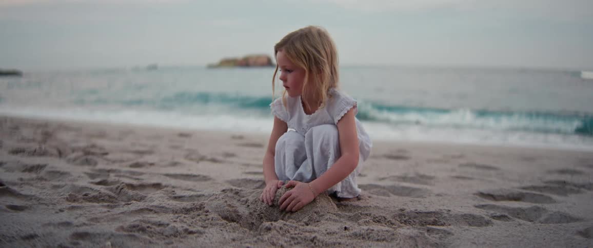A young girl wearing a light dress sits on a sandy beach and plays in the sand while ocean waves roll onto the shore under a grey sky