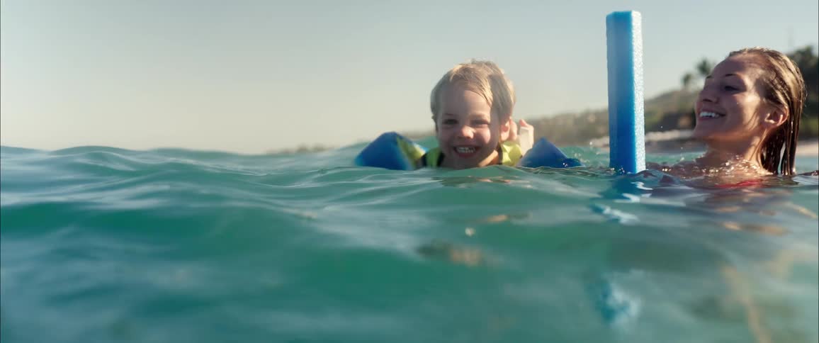 Little boy smiles as he swims in the water wearing a life jacket next to his mother in slow motion