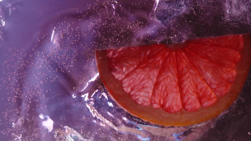 A slice of red grapefruit falls through a carbonated pink liquid and splashes against ice cubes while small bubbles and particles swirl around the fruit