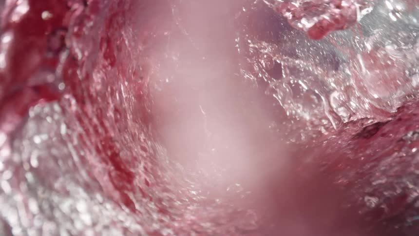 A view from inside a rotating glass shows red liquid ice cubes and citrus slices swirling together to create a colorful drink with a dynamic splashing effect