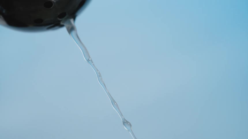 A thin stream of clear liquid pours from the bottom of a black spherical object against a solid blue background in a close view