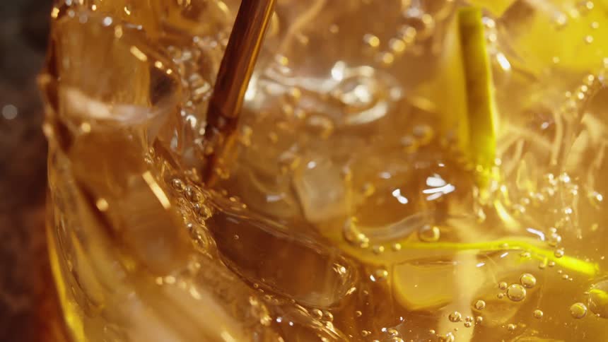 A close view inside a glass shows golden liquid and ice cubes bubbling and swirling around lime slices while a metal straw stirs the drink