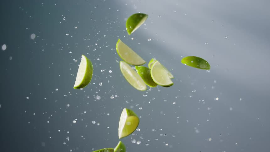 Lime wedges fall through the air filling the area with drops of juice against a grayish background