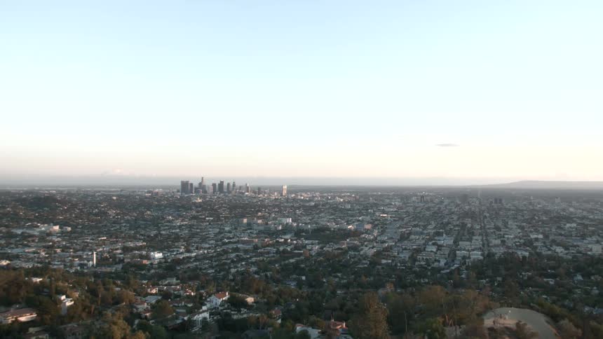 A wide aerial view shows a sprawling city with a dense skyline of tall buildings under a clear pale sky during the day