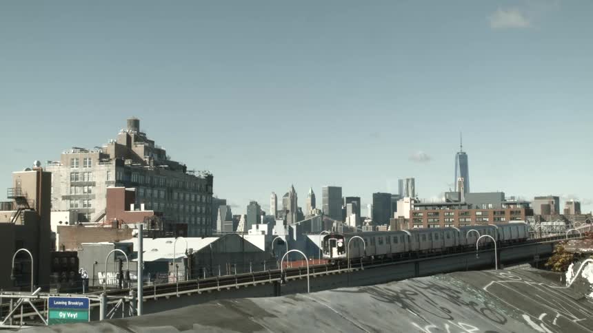 A silver subway train travels across elevated tracks in a city during the day while a sprawling urban skyline under a clear sky behind weathered brick buildings and graffiti covered walls
