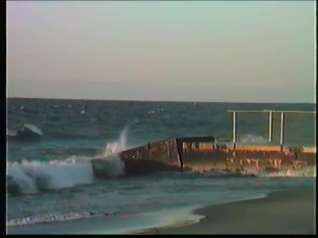 Waves crash against a weathered concrete pier and metal railing on a sandy beach while the sun illuminates the sea spray under a clear horizon