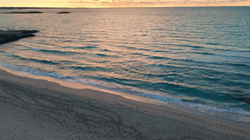 Small waves roll in and fade along a quiet sandy beach as warm sunset light spreads across the rippling water with distant land sitting low on the horizon in the background
