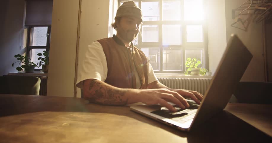 Fast push in of a tattooed creative person, wearing beanie typing on laptop in an industrial style renovated office
