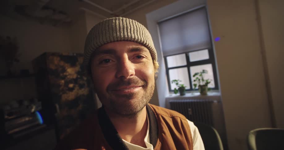A man wearing a grey beanie and a brown vest smiles and winks at the camera