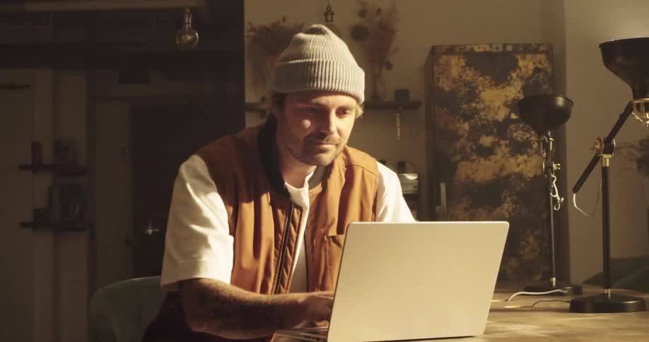 A man wearing a beanie and vest, thinking and looking up before opening his laptop and typing and starts working