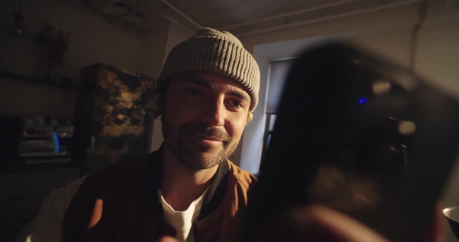 A young man sits at a desk indoors wearing a beanie smiling as he scrolls on his phone while orange sunset light streams through the window