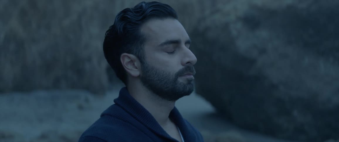 A man stands near large boulders on a beach and closes his eyes