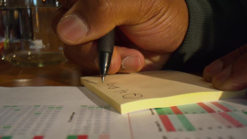 A hand uses a black pen to write q4 projects on a yellow sticky note below the sticky note a document with colorful charts and data tables is visible on the desk