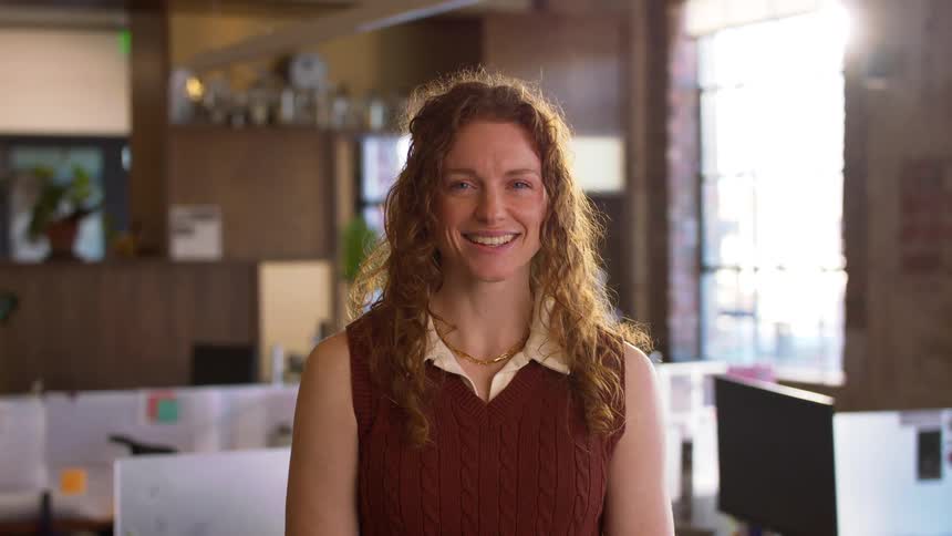 A young woman with long curly hair wearing a brown sweater vest over a light shirt stands in a modern office and smiles at the camera during the day