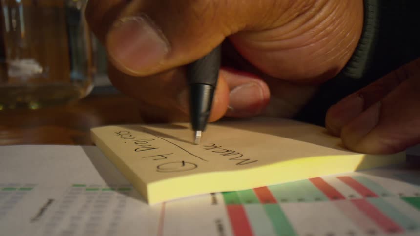 A person uses a black pen to write notes on a yellow sticky pad resting on a table next to printed documents