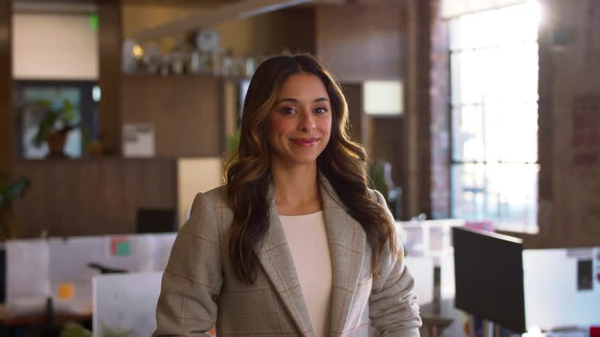 Woman in office wearing gray blazer with desks and computers in the background