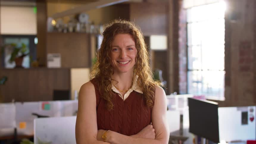 A woman with long curly red hair wearing a brown sweater vest over a white shirt smiles at the camera while standing with her arms crossed in a bright modern office with desks and a large window in the background