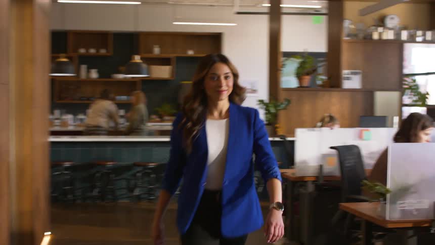 A woman wearing a blue blazer and black trousers walks through a modern office with wooden walls and a kitchen area while looking at her watch during the day