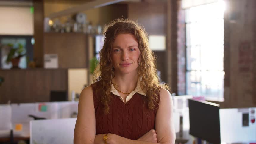 A young woman with long curly hair wearing a brown sweater vest over a light shirt stands in a modern office and smiles with her arms crossed during the day