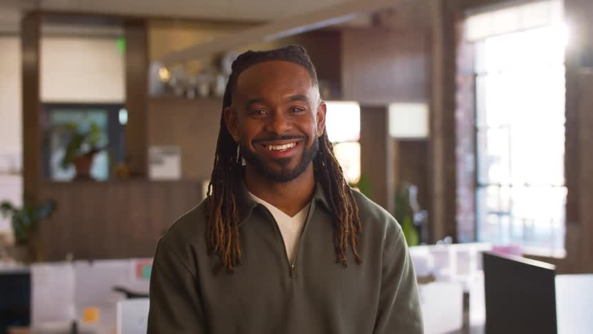 A smiling black man with long dreadlocks stands in a bright modern office wearing a green quarter zip pullover while looking directly at the camera with workstations and large windows blurred in the background