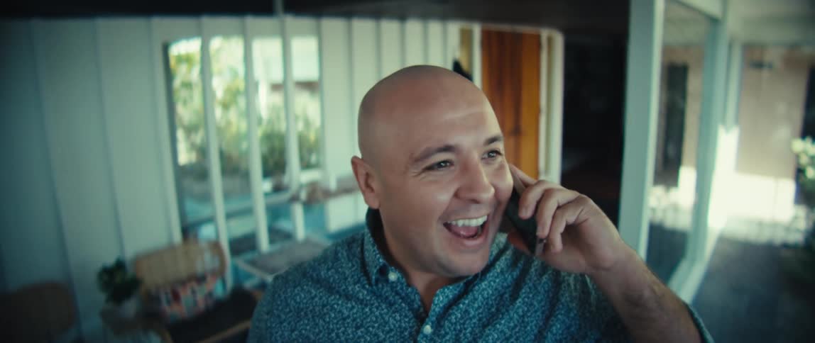 A bald man in a patterned blue shirt laughs while looking upward and then brings a phone to his earhe walks through a modern openconcept home while continuing his cheerful phone conversation