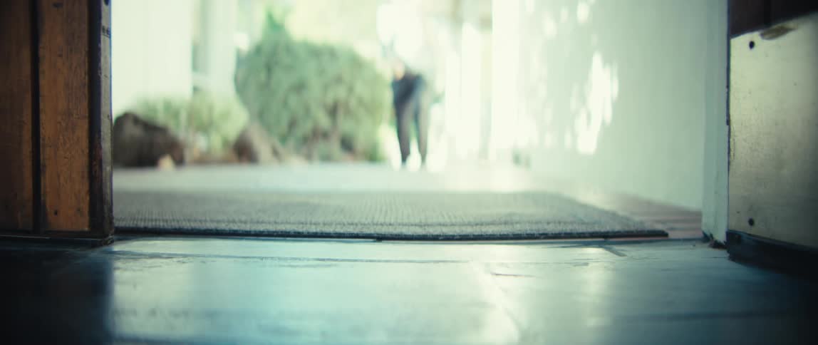 A man wearing dark trousers and shoes walks from a sunlit outdoor area through an open doorway onto a dark tiled floor