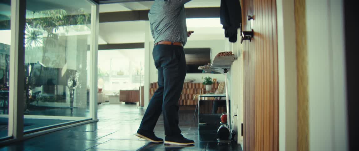 A bald man wearing a blue patterned shirt and dark trousers walks through a bright modern house while looking at his phone then picks up a jacket and opens a wooden door to exit the building