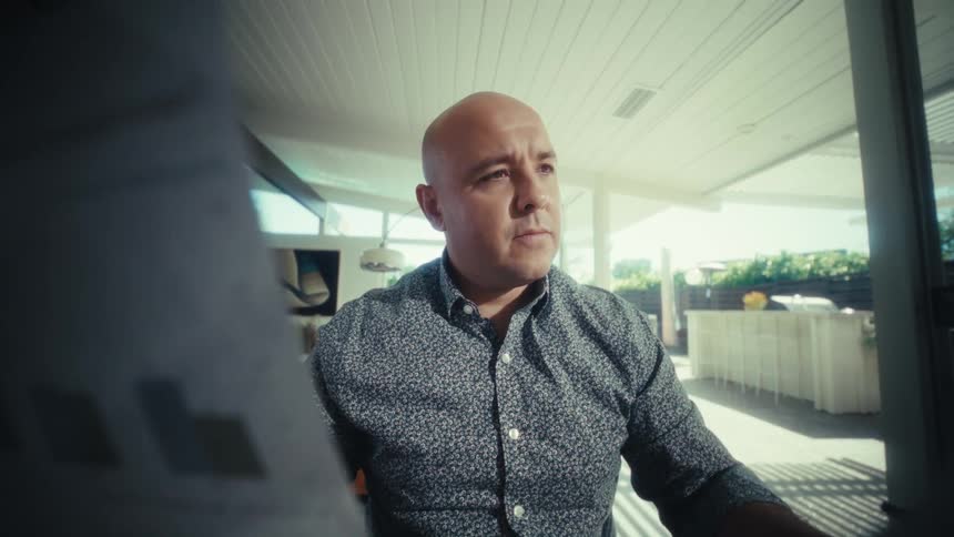 A bald man sits at a wooden dining table inside a modern house while he intensely reviews paper documents and bar charts next to a calculator laptop and glass of water with a bright patio visible in the background