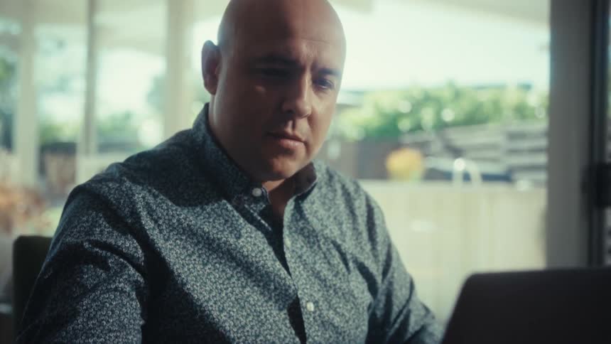 A bald man in a patterned blue shirt sits at a wooden table and looks intently at his laptop screen while working inside a bright modern house with a glass door and a sunny backyard visible in the background