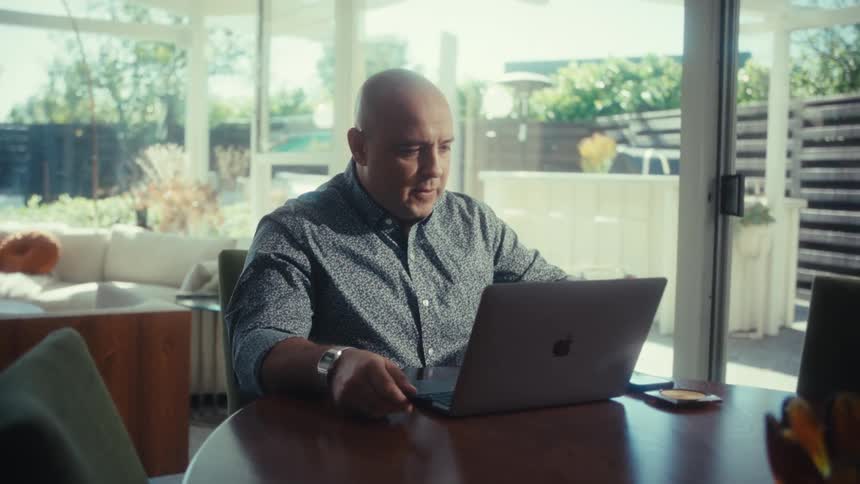 A man sits at a dining table in a bright room focused on his laptop while taking a sip from a glass of water he is dressed in a patterned grey shirt and a watch working near a large glass door that looks out onto a patio