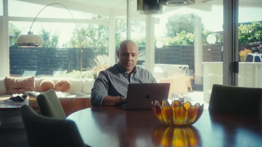 A man sitting at a table nods while looking at his laptop screen
